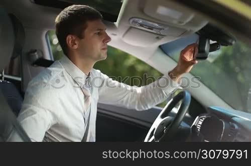 Successful businessman looking in rear-view mirror, fixing his necktie before driving car. Stylish handsome business executive in formal wear straightening his tie while sitting in driver&acute;s seat during business road trip.