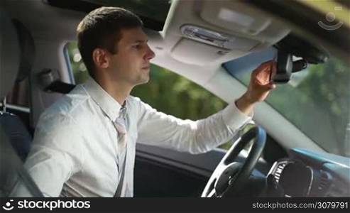 Successful businessman looking in rear-view mirror, fixing his necktie before driving car. Stylish handsome business executive in formal wear straightening his tie while sitting in driver&acute;s seat during business road trip.