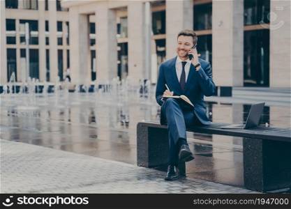 Successful businessman working outside, talking on mobile phone and smiling while sitting on bench with legs crossed, writing down information, listening to telephone call, open laptop next to him. Successful businessman talking on mobile phone while working outside, sitting on bench