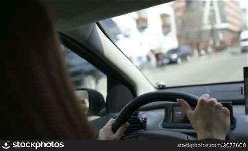 Successful businesswoman driving car through modern urban city with reflections of buildings and passing traffic. View inside out. Female hands holding steering wheel firmly while driving in city traffic closeup. Angle view.