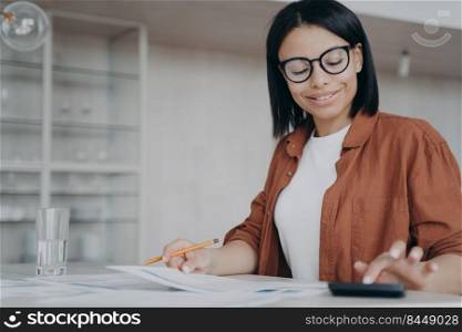 Successful businesswoman is calculating expends. Friendly young mixed race woman in glasses, entrepreneur is working remote. Workplace at home on quarantine. Finances and investment concept.. Successful mixed race businesswoman is calculating expends. Finances and investment concept.