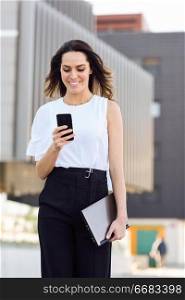 Successful businesswoman working with her smart phone and laptop outdoors. Middle-aged woman walking near business building with very careful hair.. Businesswoman working with her smart phone and laptop outdoors.