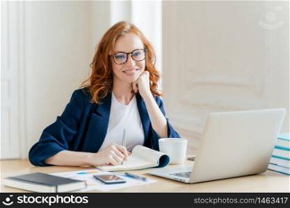 Successful economist in eyewear, checks information on laptop computer, reads information on financial website, writes in notepad and drink hot coffee, sits at workplace. Business and work concept