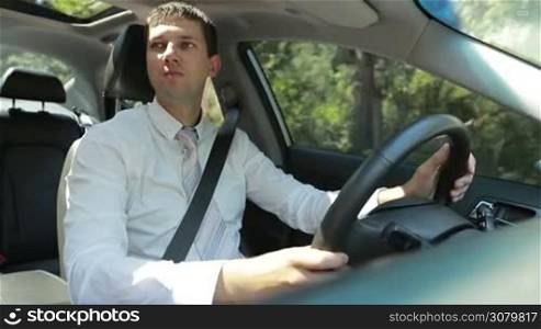 Successful entrepreneur in necktie driving car on rural road during business trip. Handsome young male driver in white shirt and tie holding steering wheel while travelling in vehicle on countryside in summer.