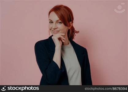 Successful europian lady with red hair isolated over pink backround, touching chin with hand and looking at camera with confident smile, happy red-hahaired female enterpreneur or business woman. Successful europian lady with red hair isolated over pink backround, touching chin with hand