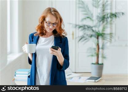 Successful female CEO reads notification on modern cell phone, has coffee break, dressed in formal clothes, starts work day from hot drink, poses over office interior, checks email box online