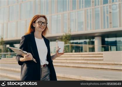Successful female entrepreneur drinks takeaway coffee carries notebook and laptop wears formal clothes spectacles goes on business meeting poses outdoor near office building. People work lifestyle. Successful female entrepreneur drinks takeaway coffee carries notebook and laptop