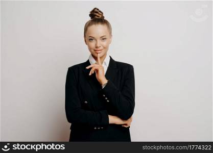 Successful female entrepreneur. Woman business owner in dark formal suit being confident and assertive with finger on lip and looking straight forward while standing isolated on grey background. Female business owner in dark suit with confident gesture