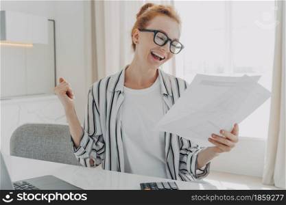 Successful female manager plans budget checks finances makes fist pump holds paper documents counts bills or taxes uses online banking service on laptop computer sits at workplace works from home. Successful female manager plans budget checks finances makes fist pump holds paper documents