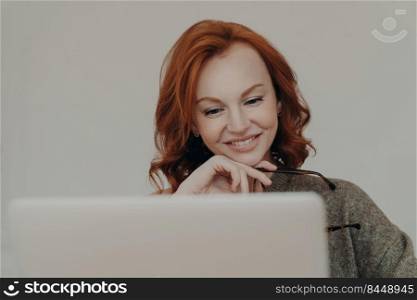Successful female student with ginger hair and pleasant smile watches interesting tutorial video or webinar, uses laptop computer for e learning, reads information online, keeps hand under chin