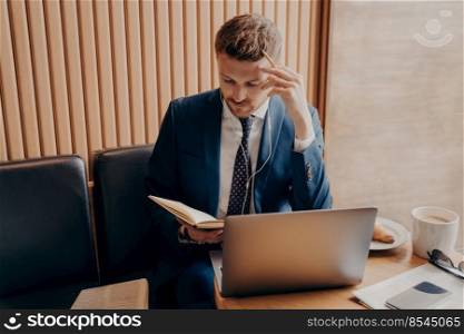 Successful financier in elegant suit with thoughtful look in earbuds and with laptop working online on business presentation while sitting in cafe over cup of latte and checking his notes in workbook. Successful financier in elegant tuxedo with thoughtful look in earbuds and with laptop