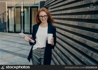 Successful ginger businesswoman standing outdoors near office with modern laptop, notebook and coffee, wearing spectacles, dressed in formal clothes, confident female employee enjoying break at work. Successful ginger businesswoman standing outdoors near office with laptop, notebook and coffee