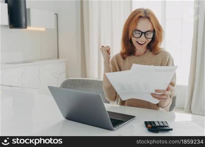 Successful ginger female freelancer checks calculations clenches fist looks happily at paper documents wears spectacles and jumper surrounded by laptop computer and calculator calculates finances. Female freelancer checks calculations clenches fist looks happily at paper documents