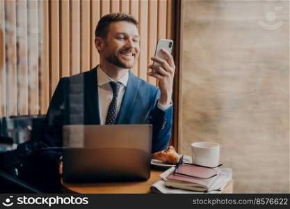 Successful handsome businessman in blue suit smiling and checking good news on smart phone while sitting in cozy cafe with laptop and working remotely, documents and croissant in front of him on table. Successful businessman chatting on smart phone while working in cafe