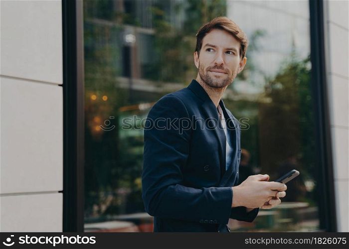 Successful handsome businessman with stubble messaging his employer about project details, holding smartphone in his hands while standing next to building with big window reflecting urban landscape. Successful handsome businessman messaging to client on smartphone while standing next to building