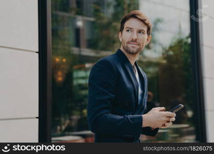 Successful handsome businessman with stubble messaging his employer about project details, holding smartphone in his hands while standing next to building with big window reflecting urban landscape. Successful handsome businessman messaging to client on smartphone while standing next to building