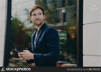 Successful handsome businessman with stubble messaging his employer about project details, holding smartphone in his hands while standing next to building with big window reflecting urban landscape. Successful handsome businessman messaging to client on smartphone while standing next to building