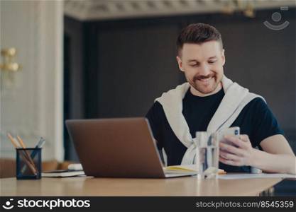 Successful handsome male freelancer in casual wear looking at smartphone and smiling while reading messages or good news, sitting at his workplace in front of laptop at home. Business and work concept. Pleasant freelancer using mobile phone while working remotely from home