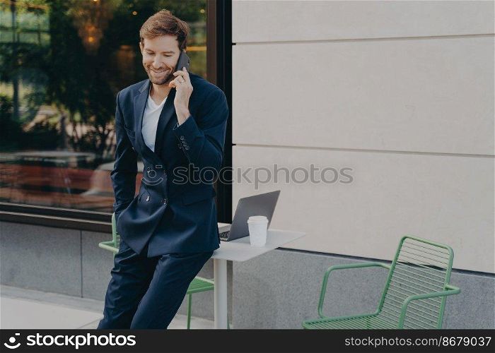 Successful handsome male trader discussing stock prices on while leaning on sidewalk cafe table, enjoying being self employed, happy after hearing good news about his investments. Successful handsome male trader discussing stock prices on while leaning on sidewalk cafe table