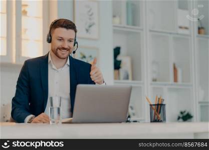 Successful happy businessman in suit holding thumb up and looking at laptop screen with smile while communicating online with colleague, working remotely from home. Distant job concept. Happy young man showing thumb up while having video conference