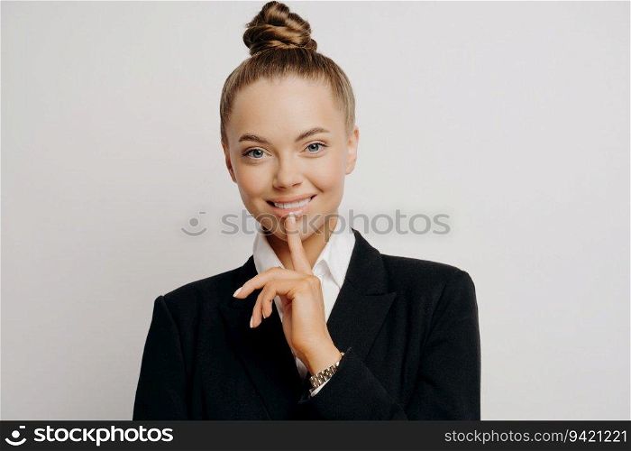 Successful lawyer in dark suit, hair in bun, smiling, finger on lips, hearing good news about opportunities, grey wall.