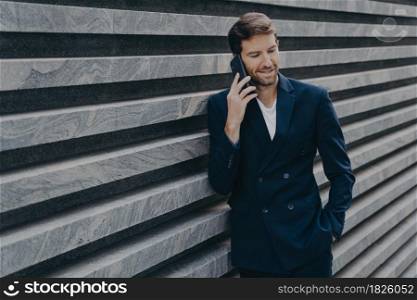 Successful male lawyer makes consultation with partner keeps hand in pocket of black suit arranges meeting via smartphone poses near marble wall copy space. People business and technology concept. Successful male lawyer makes consultation talks via smartphone