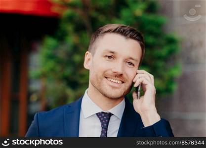 Successful male office worker has conversation about business affairs via cell phone, dressed in formal clothes, going to have lunch break at cafe, poses outside, has positive smile on face.