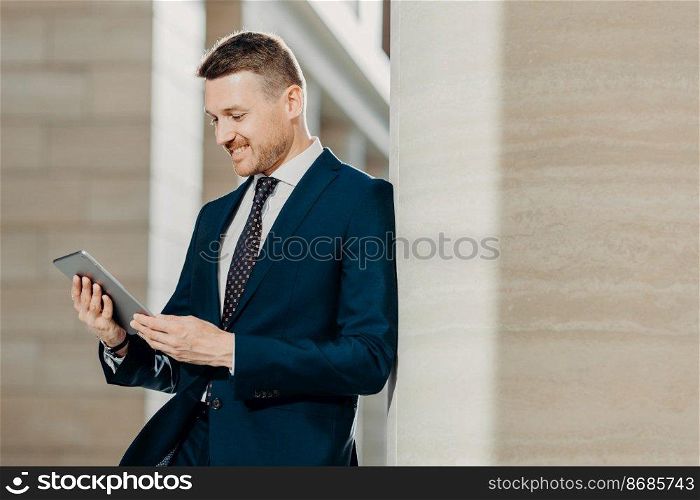 Successful male office worker in black suit uses modern tablet computer for distance work, connected to wireless internet, poses in modern interior, concentrated into screen. Technology concept