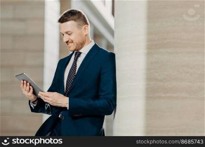 Successful male office worker in black suit uses modern tablet computer for distance work, connected to wireless internet, poses in modern interior, concentrated into screen. Technology concept