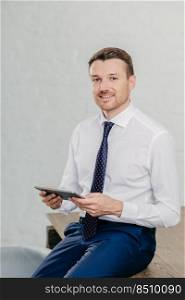 Successful owner of financial company wears elegant clothes, reads notification or chatts with friends on modern touch pad, has gentle smile, looks confidently at camera. People and technology concept