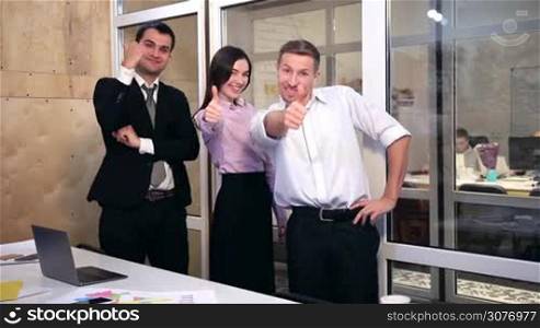 Successful positive business team giving thumbs up sign and smiling. Happy group of business people standing at the table in modern office and giving thumbs up.