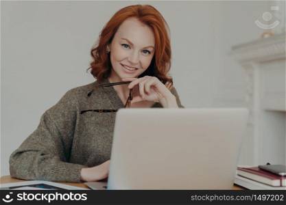 Successful professional businesswoman with red hair works remotely on laptop computer, reads article about business affairs online, sits at desktop, uses modern technologies, prepares financial report