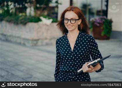 Successful redhead female office worker returns from work poses outside with organizer modern gadgets wears spectacles black polka dot dress focused into distance strolls at street. Business people. Female office worker poses outside with organizer modern gadgets