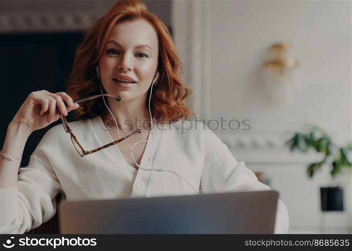 Successful redhead woman entrepreneur ready to conduct online negotiations with coworkers, holds spectacles, sits in front of opened laptop, uses earphones, searches needed information in internet