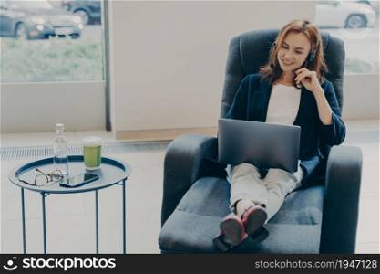 Successful redhead woman entrepreneur talking online with coworkers, sitting on armchair in relaxed pose in front of opened laptop and using headset, working distantly at home. Freelance concept. Successful redhead woman entrepreneur talking online with coworkers while working remotely at home