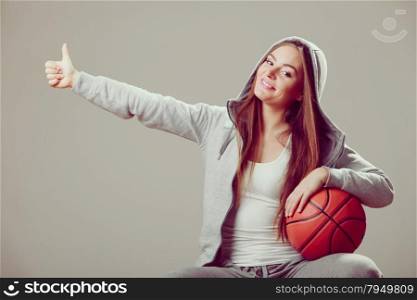 Successful sporty teenager girl wearing hooded sweatshirt holding basketball with thumb up gesture. Teen sport.