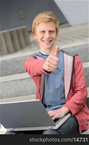 Successful teenage student show thumb-up sitting with laptop on steps