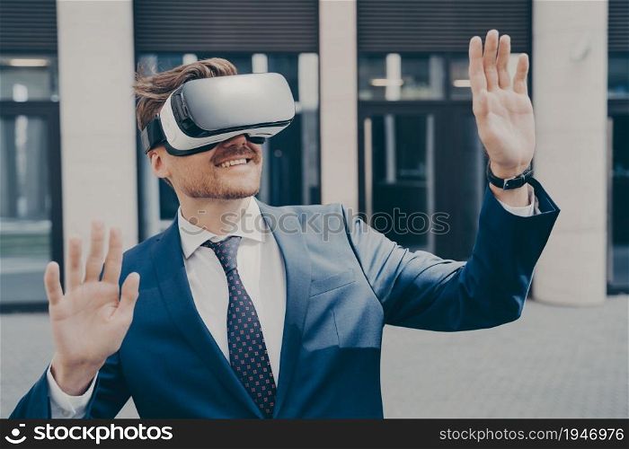 Successful video game publisher wearing black and white VR headset, amazed bearded businessman trying out new immersive virtual reality game, playing outside, building in blurred background. Successful video game publisher wearing black and white VR headset while standing outside