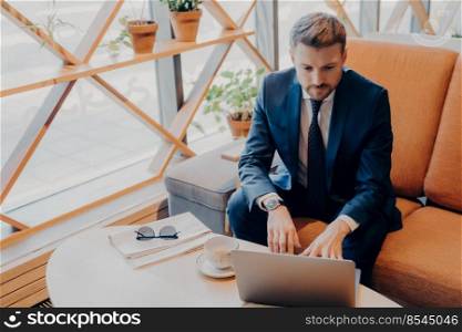Successful well-groomed financier in stylish suit with trimmed beard sits in business centre lobby over cup of tea while waiting for meeting and takes final notes on his notebook computer online. Successful well-groomed financier in stylish suit with trimmed beard sits in lobby