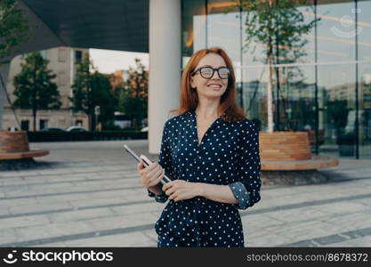 Successful woman entrepre≠ur carrying laptop notebook whi≤standing on city street, looking aside with smi≤, smiling fema≤freelancer with digital device in hand enjoying beautiful day outside. Successful happy woman entrepre≠ur carrying laptop notebook whi≤standing on city street