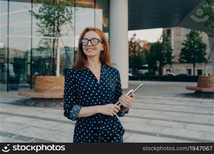 Successful woman entrepreneur carrying laptop notebook while standing on city street, looking aside with smile, smiling female freelancer with digital device in hand enjoying beautiful day outside. Successful happy woman entrepreneur carrying laptop notebook while standing on city street