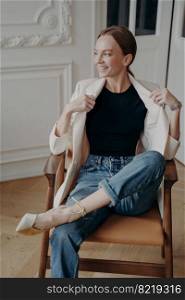 Successful woman is sitting in vintage armchair with her legs crossed. Portrait of stylish young adult european lady in white jacket posing in studio. Concept of elegance and confidence.. Successful woman is sitting in armchair with her legs crossed. Stylish young adult european lady.