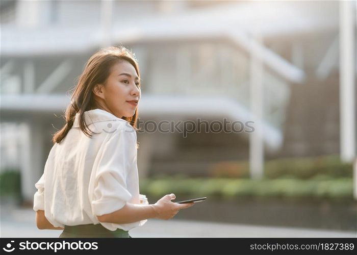 Successful young Asia businesswoman in fashion office clothes holding disposable paper cup of hot drink and using smart phone while walking outdoors in urban modern city. Business on the go concept.