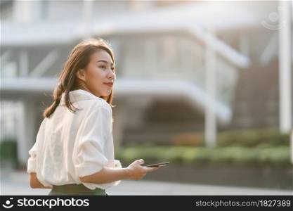 Successful young Asia businesswoman in fashion office clothes holding disposable paper cup of hot drink and using smart phone while walking outdoors in urban modern city. Business on the go concept.