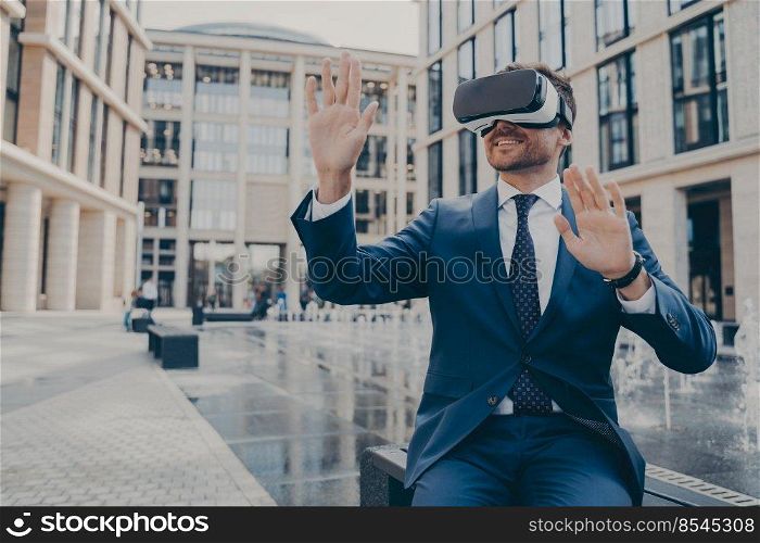 Successful young businessman dressed formally viewing future project in virtual reality using VR goggles while sitting on bench, fountain and buildings on blurred background. Successful young businessman in formal suit viewing future project in virtual reality