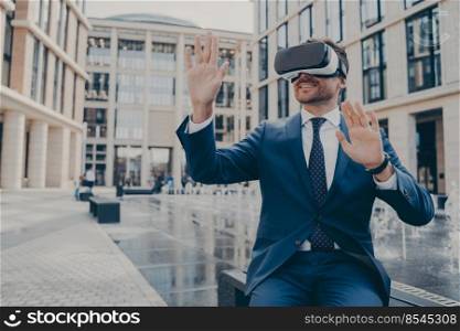 Successful young businessman dressed formally viewing future project in virtual reality using VR goggles while sitting on bench, fountain and buildings on blurred background. Successful young businessman in formal suit viewing future project in virtual reality