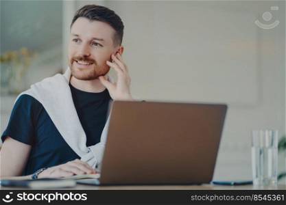 Successful young businessman wearing earphones looking aside, feeling happy and smiling after online meeting. Freelancer thinking about positive outcome of conversation while working on laptop at home. Cheerul man working remotely on laptop at home, having online meeting