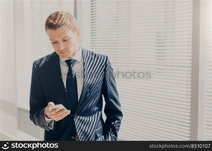 Successful young entrepreneur in expensive suit, holds mobile phone, stands in office, against window in cabinet, chats online, reads news about finances, connceted to wireless internet at work