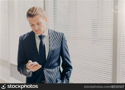 Successful young entrepreneur in expensive suit, holds mobile phone, stands in office, against window in cabinet, chats online, reads news about finances, connceted to wireless internet at work