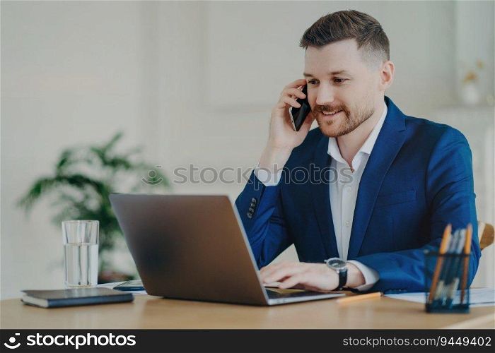 Successful young entrepreneur in formal suit smiles, talks on phone, discusses business with partner in modern office.